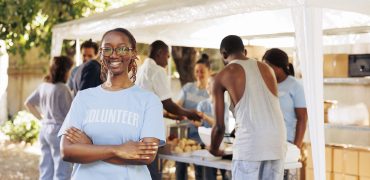 Young black woman with glasses stands outdoors, arms crossed, looking at the camera. Diverse group of volunteers supports a non-profit program dedicated to hunger relief and helping needy individuals.